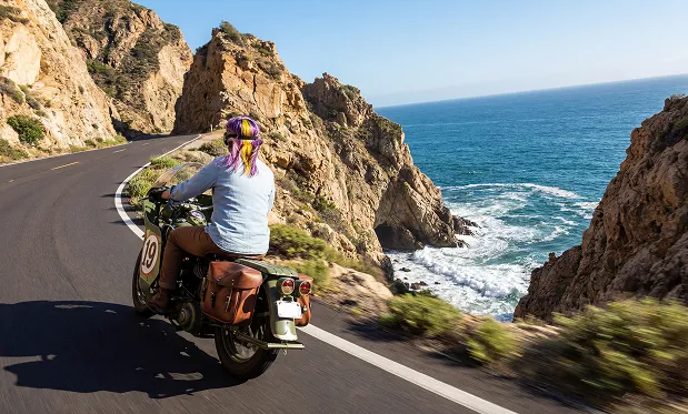 Eine Person fährt ein grünes Motorrad auf einer kurvenreichen Küstenstraße, das dunkelblaue Meer zieht sich neben steilen Klippen entlang. Das Bild zeigt eine dynamische...