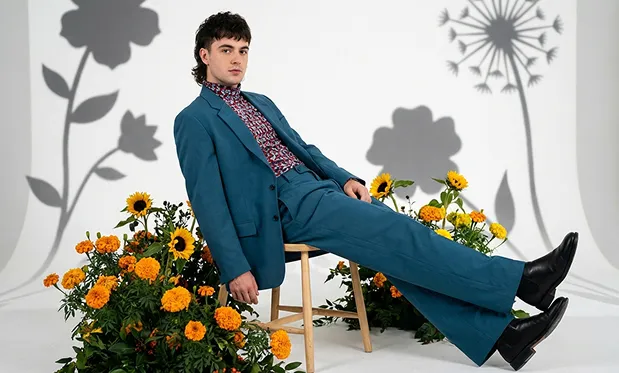 一名身穿藍色西裝及圖案襯衫的男子，坐在小凳子上，四周環繞著鮮艷的橙色和黃色花卉，背景為純白色。圖像展現...